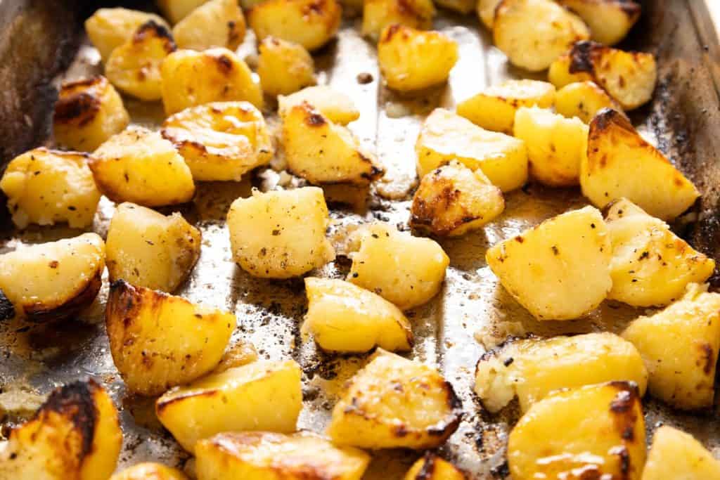 Golden roasted potato chunks on a metal baking tray, seasoned with herbs and lightly browned, with a crispy texture visible on some pieces—these are classic Greek Lemon Potatoes, bursting with bright citrus flavor and aromatic spices.