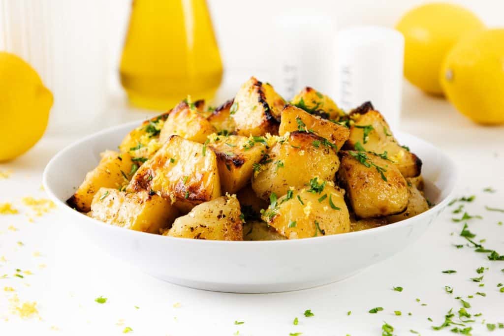 A white bowl filled with golden, roasted Greek Lemon Potatoes garnished with chopped parsley. Lemons, oil, and salt and pepper shakers are blurred in the background on a white surface.