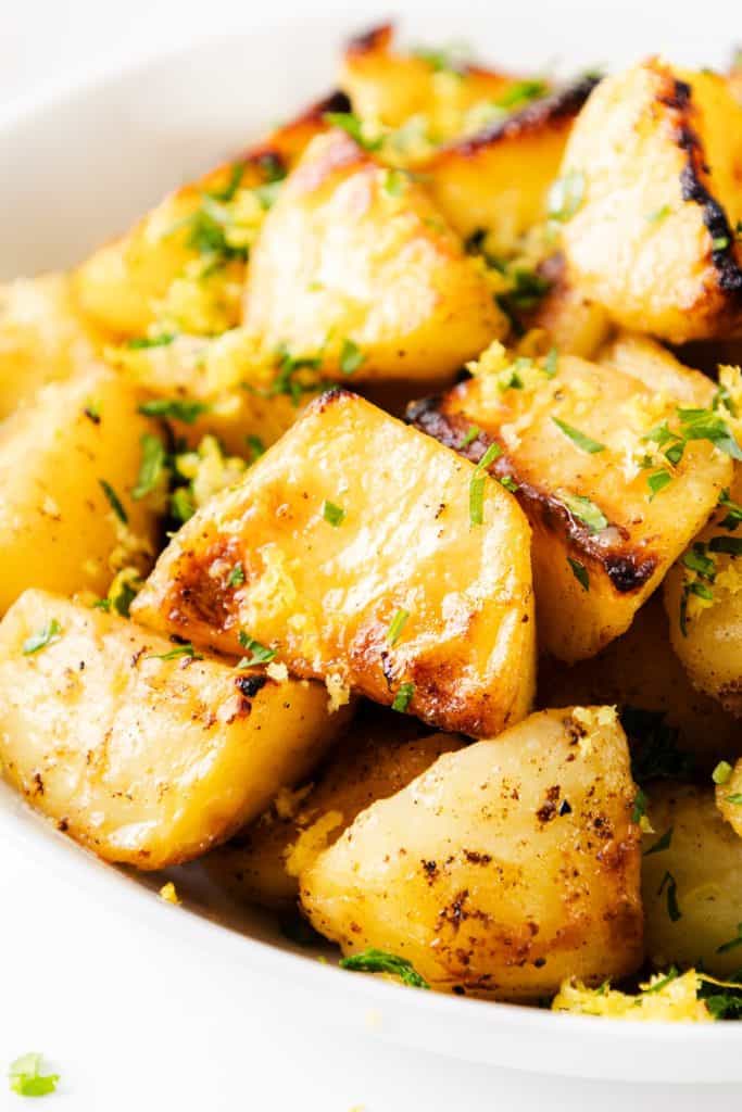 Close-up of golden, roasted Greek Lemon Potatoes garnished with chopped herbs and grated zest, served in a white bowl.