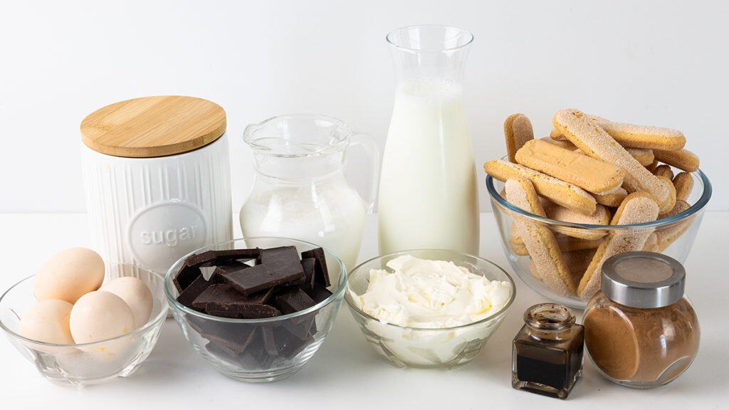 A variety of Hot Chocolate Tiramisu ingredients on a white surface: eggs, sugar, chocolate, mascarpone, ladyfingers, milk, cream, coffee, and cocoa powder arranged in bowls and jars.