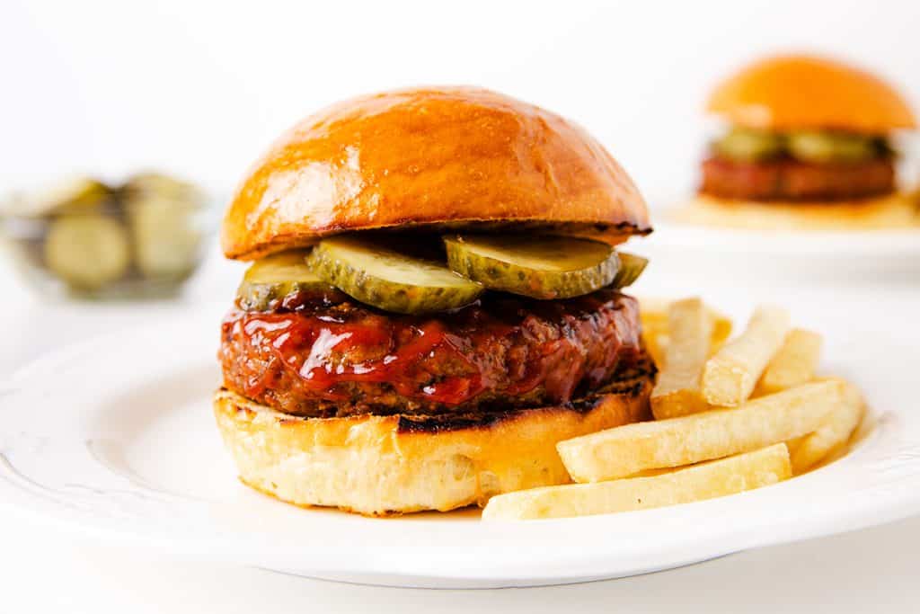 A juicy meatloaf burger with pickles and barbecue sauce in a shiny bun is served with French fries on a white plate. In the background, another burger and a jar of pickles are visible.