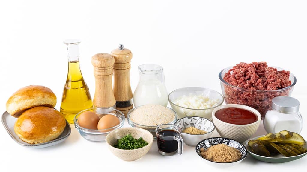 A variety of burger ingredients for Meatloaf Burgers—including buns, eggs, ground beef, chopped onions, breadcrumbs, parsley, pickles, ketchup, and spices—are all arranged on a white surface.