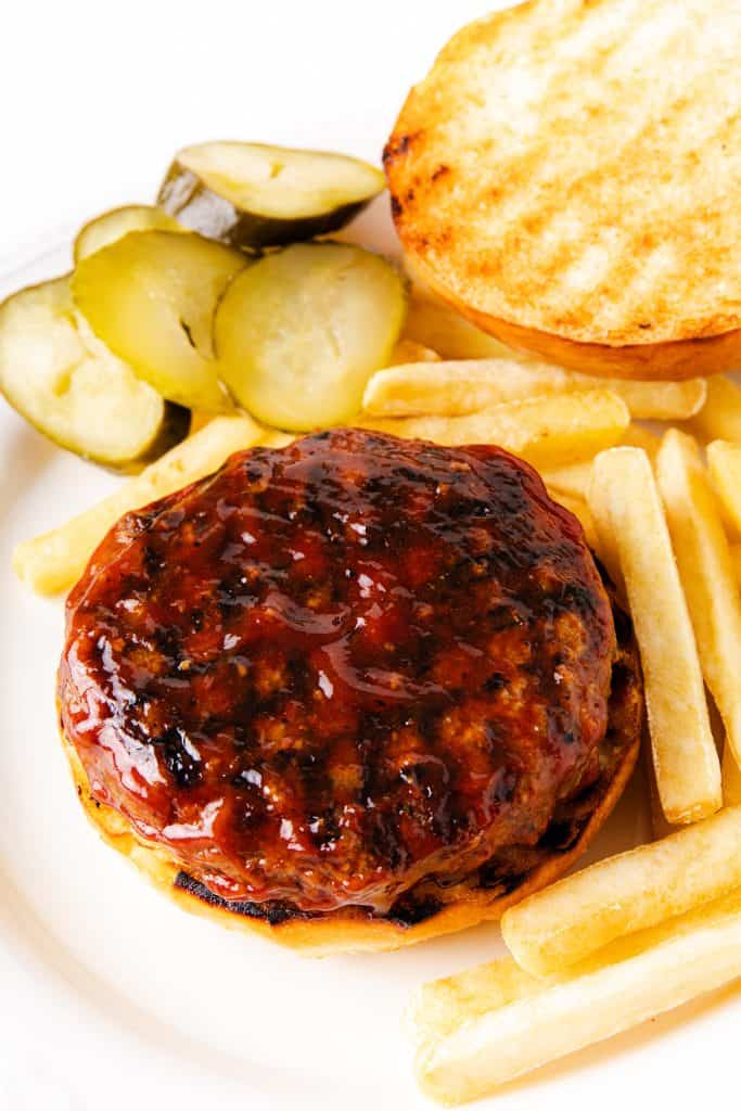 An open-faced Meatloaf Burger with a glazed beef patty on a toasted bun, served alongside French fries and sliced pickles on a white plate.