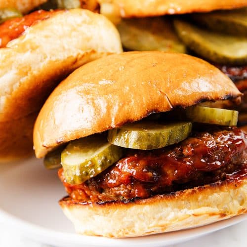 A close-up of a mouthwatering Meatloaf Burger with a shiny bun, juicy beef patty, barbecue sauce, and sliced pickles, served on a white plate with another burger in the background.