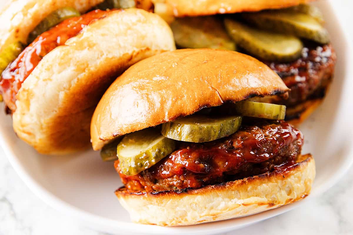 A close-up of a mouthwatering Meatloaf Burger with a shiny bun, juicy beef patty, barbecue sauce, and sliced pickles, served on a white plate with another burger in the background.