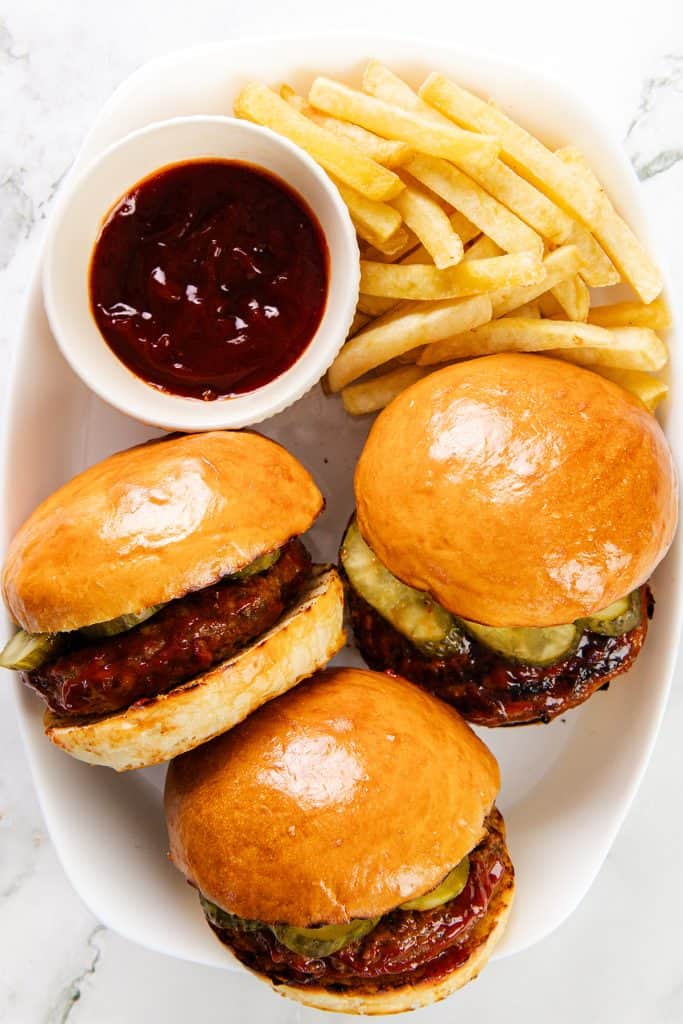 A plate with three Meatloaf Burgers sliders slathered in barbecue sauce and topped with pickles, served alongside golden French fries and a small bowl of barbecue sauce on the side.