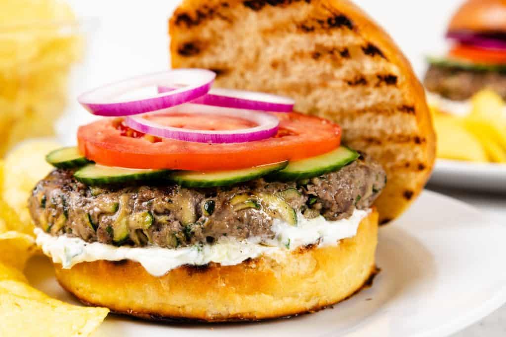 A Mediterranean-inspired grilled lamb burger with lettuce, tomato, cucumber, red onion, and a creamy white sauce on a toasted bun, served on a white plate with potato chips on the side.
