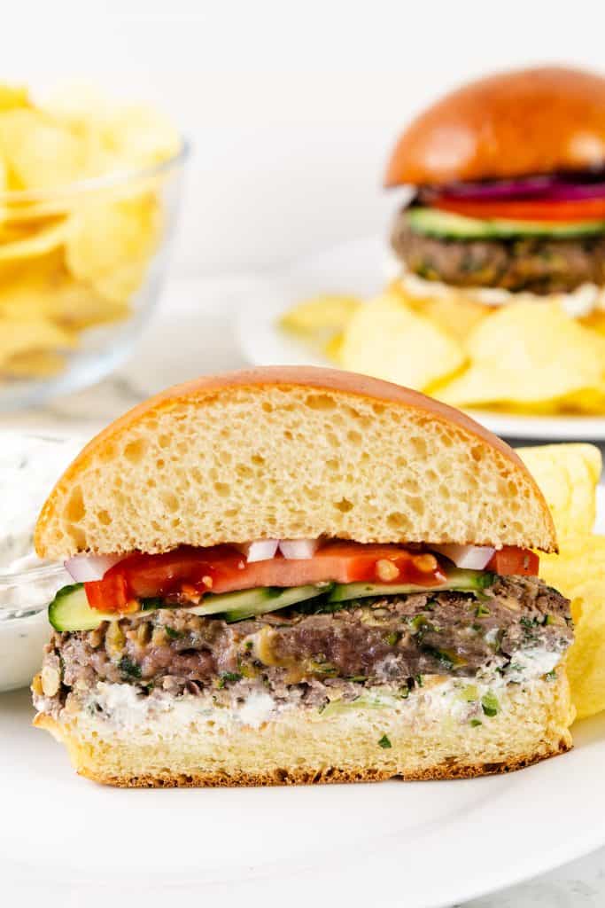 A close-up of a half black bean burger on a bun with onions, tomato, cucumber, and sauce, served alongside potato chips. Another burger and chips are blurred in the background&mdash;a vibrant scene reminiscent of Mediterranean flavors often found in lamb burgers.