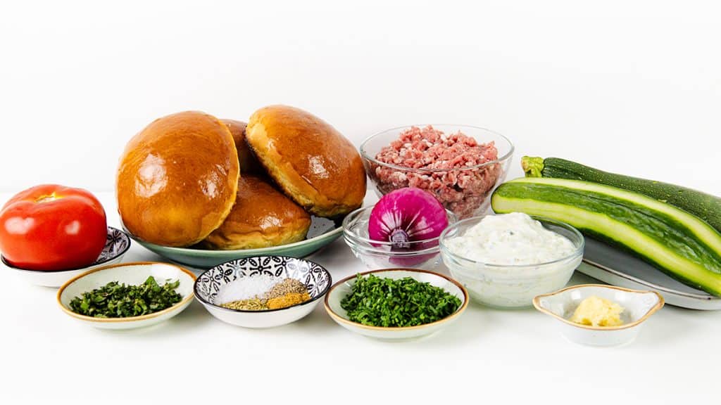 Three burger buns, minced meat, a tomato, a red onion, two zucchinis, bowls of herbs and spices, tzatziki, mustard, and chopped greens arranged on a white surface&mdash;perfect for creating delicious Mediterranean Lamb Burgers.