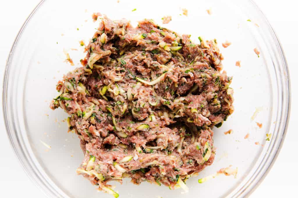 Ground lamb meat mixture with shredded vegetables, likely zucchini, in a clear glass bowl on a white surface&mdash;this Mediterranean-inspired blend is ready to be formed into delicious lamb burgers or meatballs.