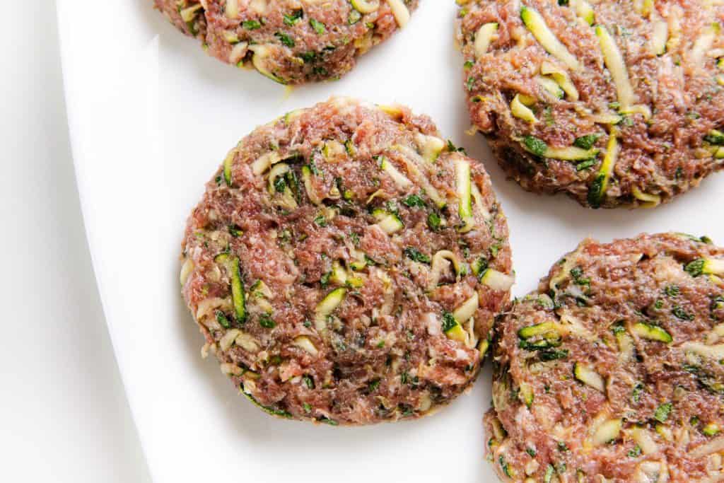 Raw, uncooked lamb burger patties mixed with shredded vegetables, likely zucchini, arranged on a white plate. The patties have a coarse texture with visible green and white veggie pieces, giving them a fresh Mediterranean flair.