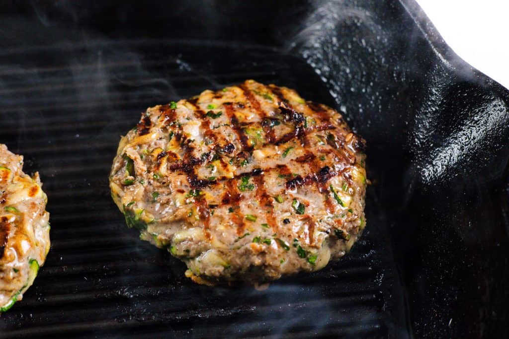 A grilled Mediterranean Lamb burger patty with visible grill marks cooks on a black grill pan, with steam rising around it.