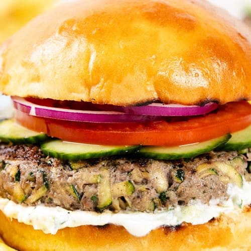 A close-up of a Mediterranean Lamb burger with lettuce, tomato, cucumber, red onion, and sauce in a bun, served with potato chips on a white plate.