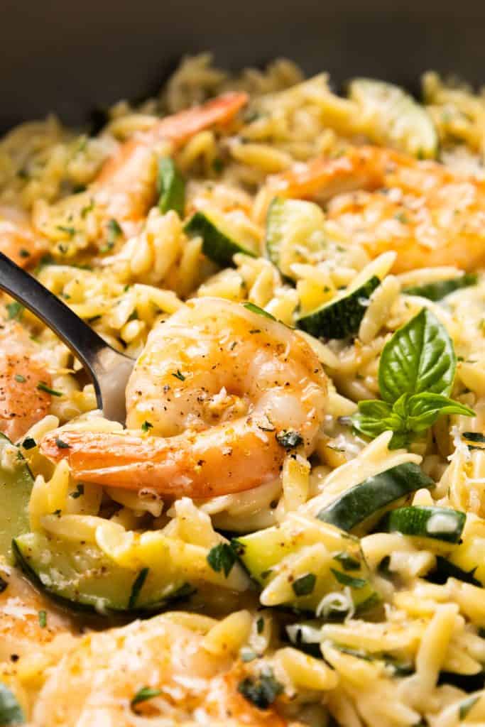 Close-up of cooked shrimp on a bed of creamy one-pan orzo with zucchini slices, garnished with herbs and a fresh basil leaf. A fork is picking up one shrimp, highlighting the flavorful and well-seasoned dish.