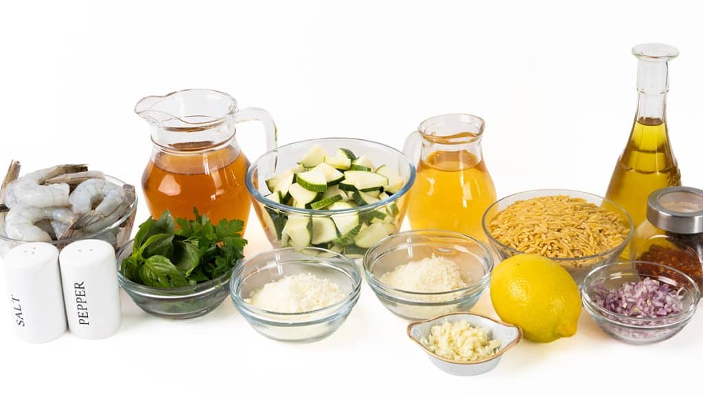 A variety of ingredients are arranged on a white surface, ready for a delicious One-Pan Orzo creation. Fresh shrimp, vibrant zucchini, herbs, lemon, broth, oil, orzo pasta, grated cheese, chopped onions, and seasonings are organized in bowls and containers.