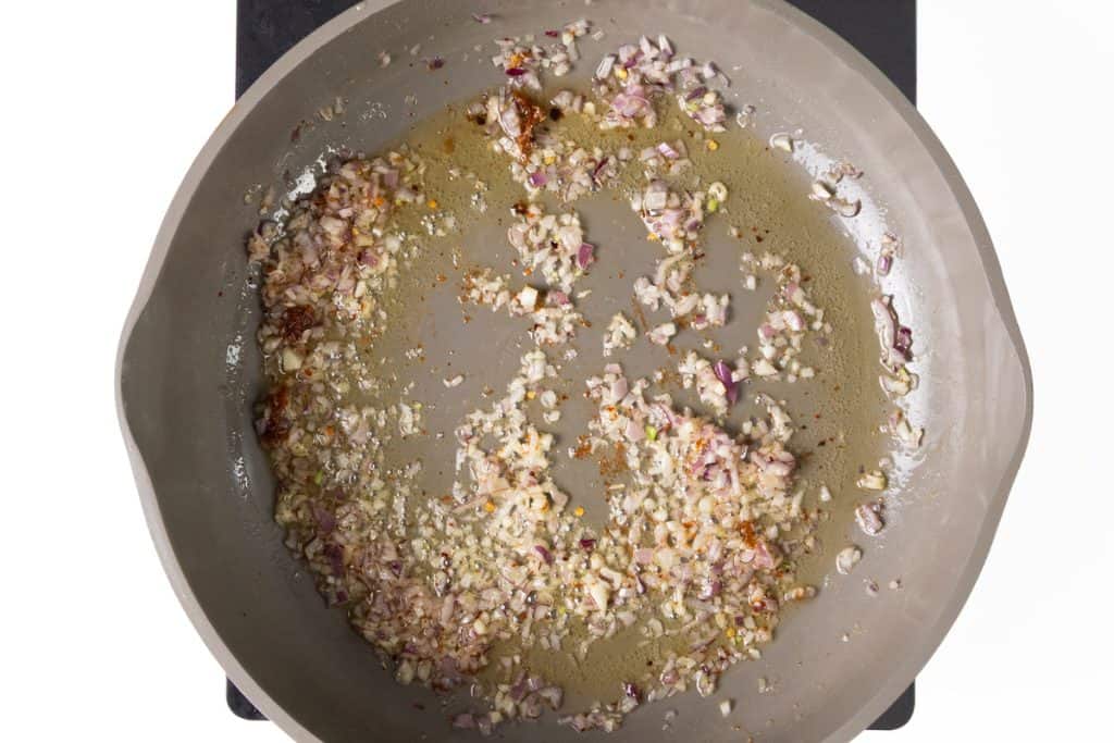Chopped onions and garlic are being sautéed in oil in a skillet, beginning to brown and soften alongside sliced zucchini, on a stovetop with a white background.