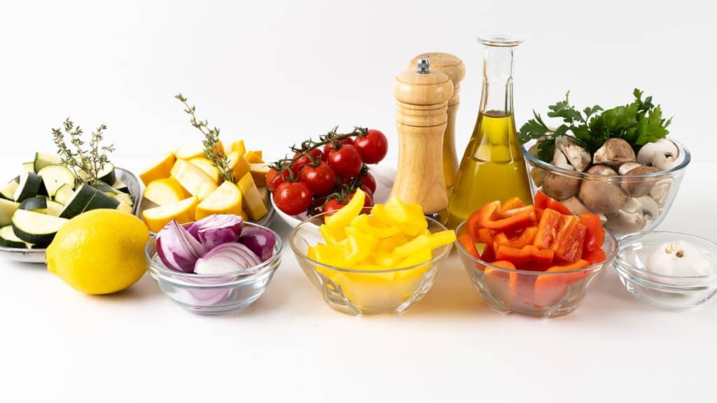 Various fresh summer vegetables, including zucchini, yellow squash, tomatoes, mushrooms, bell peppers, red onion, and a lemon, sit alongside garlic, parsley, olive oil, and salt and pepper shakers on a white background—perfect for creating flavorful roasted vegetables with a garlic herb finish.