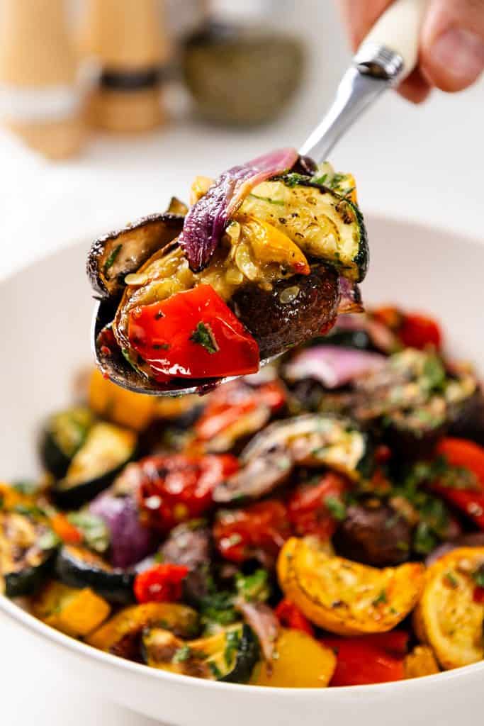 A close-up of a spoonful of colorful Garlic-Herb Roasted Summer Vegetables, including zucchini, red peppers, onions, and eggplant, held above a bowl brimming with more roasted vegetables.