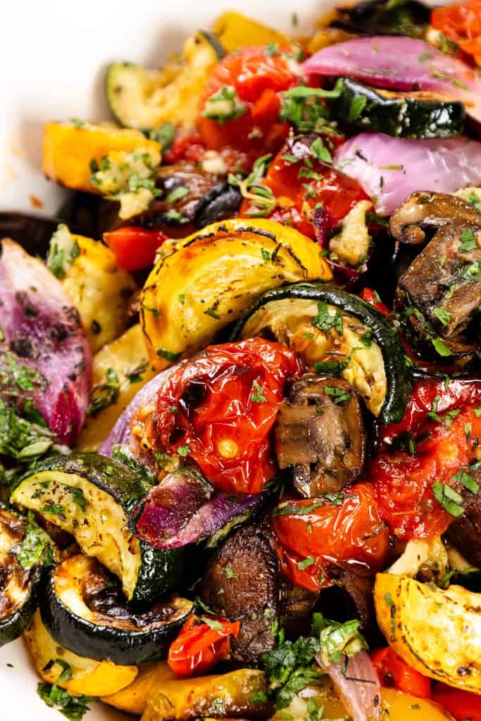 A close-up of colorful garlic-herb roasted vegetables including zucchini, tomatoes, red onions, yellow squash, and mushrooms, garnished with chopped herbs.