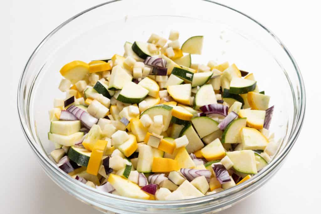 A glass bowl filled with chopped vegetables, including yellow squash, zucchini, red onion, and white root vegetables, sits on a white surface&mdash;perfect for tossing into your favorite summer recipes or creating a vibrant Summer Vegetable Pappardelle.