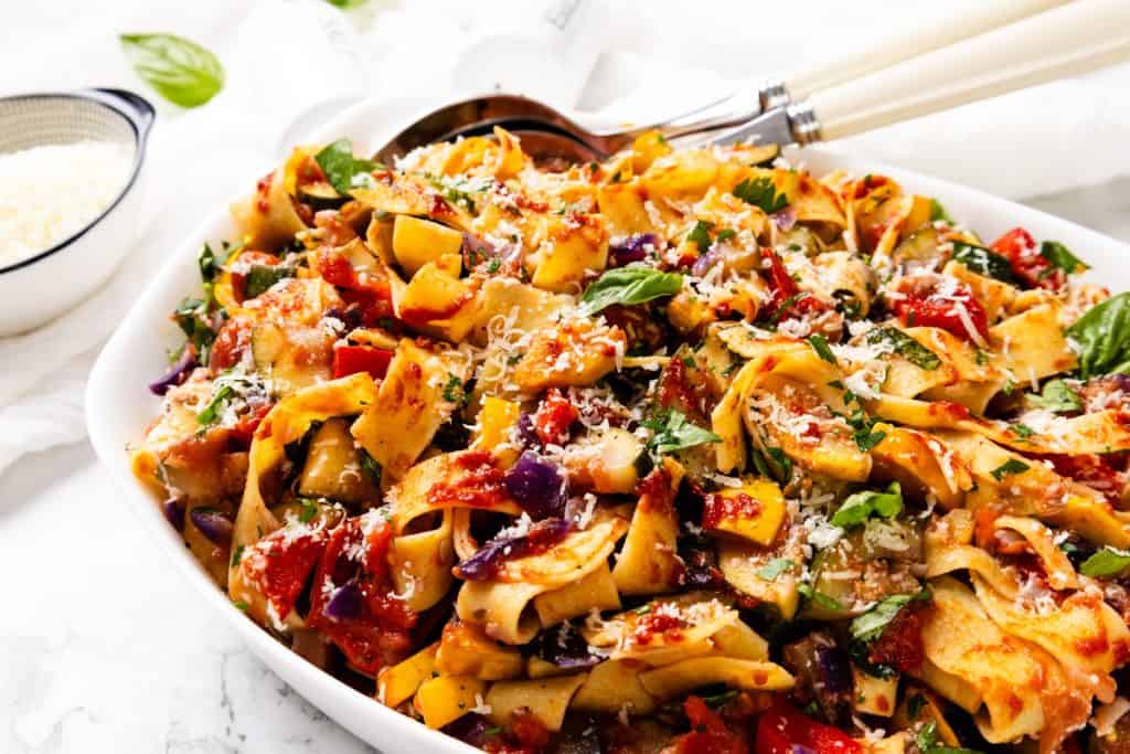 A close-up of a white dish filled with wide pappardelle noodles, vibrant summer vegetables, tomato sauce, fresh basil, and grated cheese, with serving utensils resting on the side.