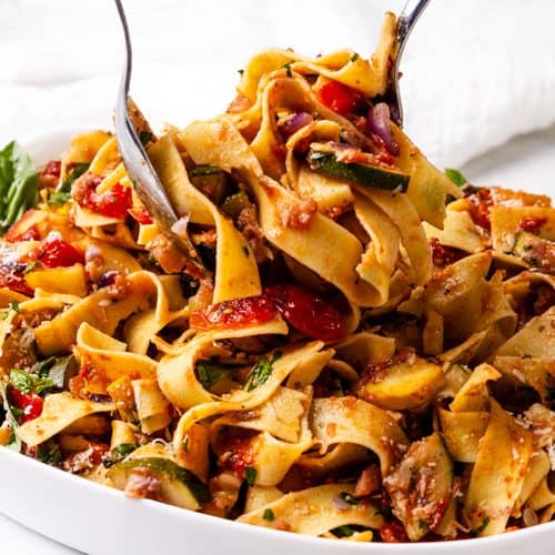 A bowl of wide pappardelle noodles mixed with tomatoes, zucchini, herbs, and sauce is being served with tongs on a white background&mdash;an inviting plate of Summer Vegetable Pappardelle.