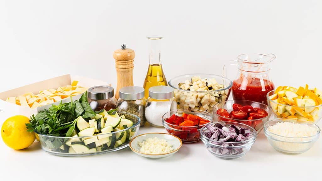 Assorted fresh vegetables, seasonings, oil, herbs, and grated cheese are arranged in bowls and jars on a white surface, ready for cooking a vibrant Summer Vegetable Pappardelle.