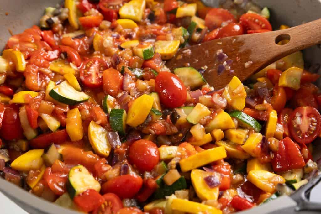 Colorful chopped vegetables, including tomatoes, zucchini, and yellow squash, are being saut&eacute;ed in a pan with diced onions to create the flavorful base for a Summer Vegetable Pappardelle. A wooden spoon is stirring the vibrant mixture, bringing the fresh tastes of summer together in every bite.