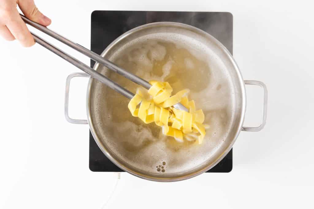 A hand uses tongs to lift uncooked summer vegetable pappardelle above a boiling pot of water on a stovetop. Steam rises from the pot as the pasta begins to cook.