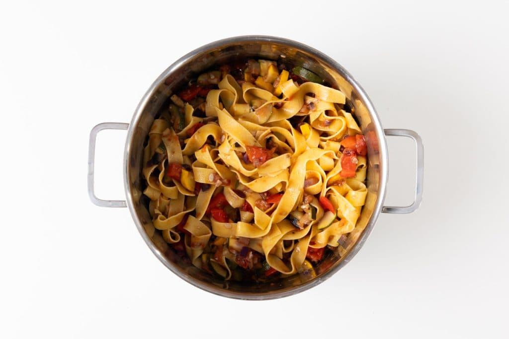 A stainless steel pot filled with cooked fettuccine pasta, mixed with diced tomatoes, bell peppers, zucchini, and mushrooms&mdash;an inviting vegetable pasta perfect for summer recipes&mdash;shown from above on a white background.