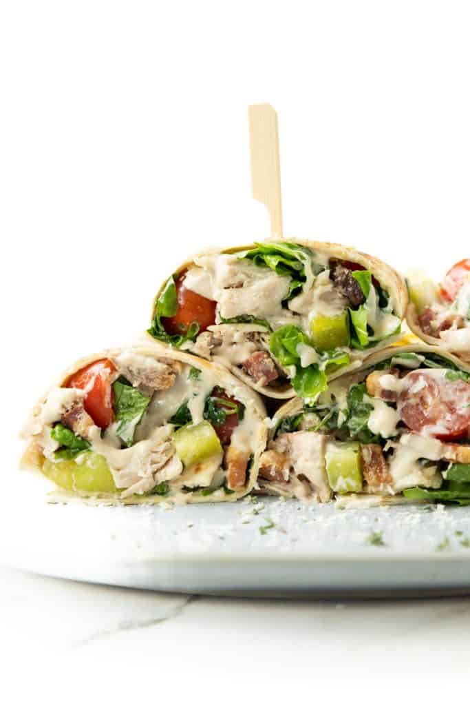 A close-up of a Chicken Caesar Wrap cut in half, filled with lettuce, tomato, cucumber, grilled chicken, and drizzled with creamy dressing, held together with a wooden pick on a white plate.