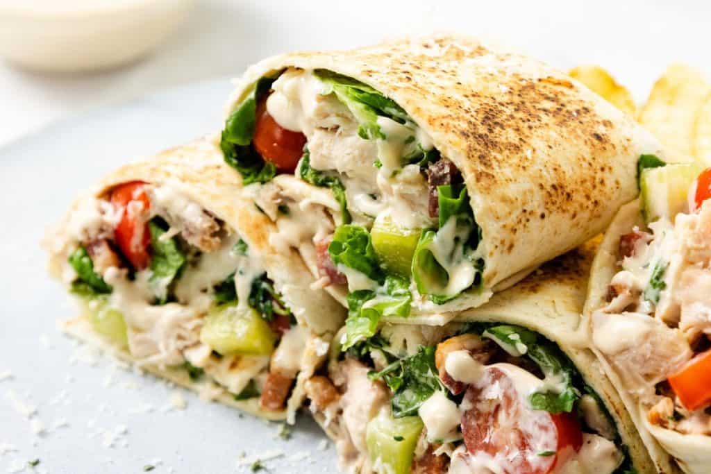 Close-up of a Chicken Caesar Wrap cut in half, filled with lettuce, tomatoes, cucumbers, shredded meat, and drizzled with creamy sauce, served on a plate.