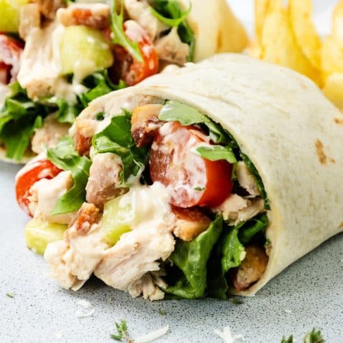 A close-up of a Chicken Caesar Wrap filled with lettuce, cherry tomatoes, cucumber, and sauce, served with potato chips on the side.
