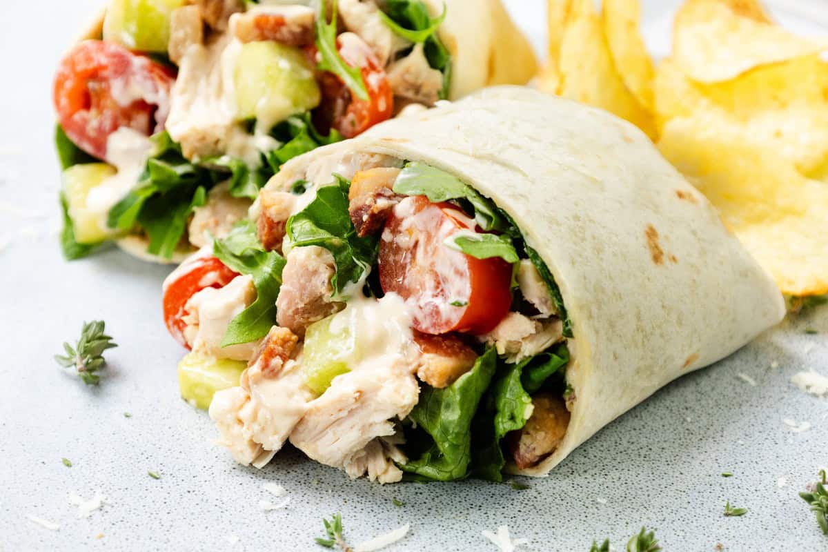 A close-up of a Chicken Caesar Wrap filled with lettuce, cherry tomatoes, cucumber, and sauce, served with potato chips on the side.