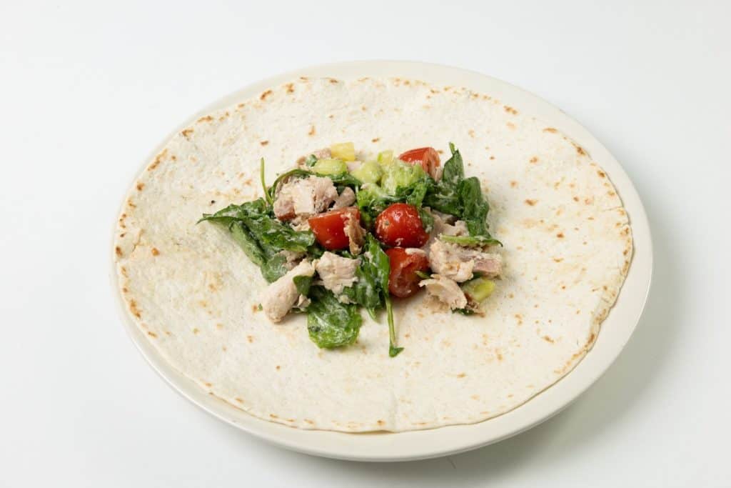 An open tortilla on a white plate topped with a mixture of tuna, cherry tomatoes, lettuce, and cucumber, reminiscent of the freshness found in a Chicken Caesar Wrap, all set against a white background.