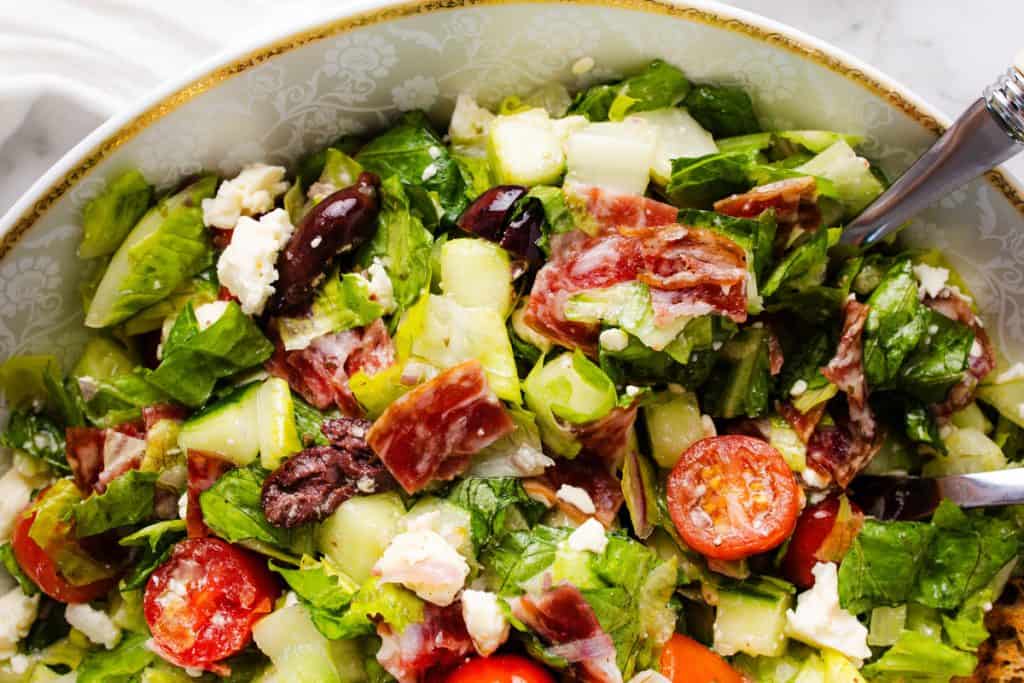 Chopped Salad with crisp lettuce, cherry tomatoes, cucumber, Kalamata olives, feta cheese, and slices of cured meat, served in a decorative bowl with a silver utensil visible on the side.