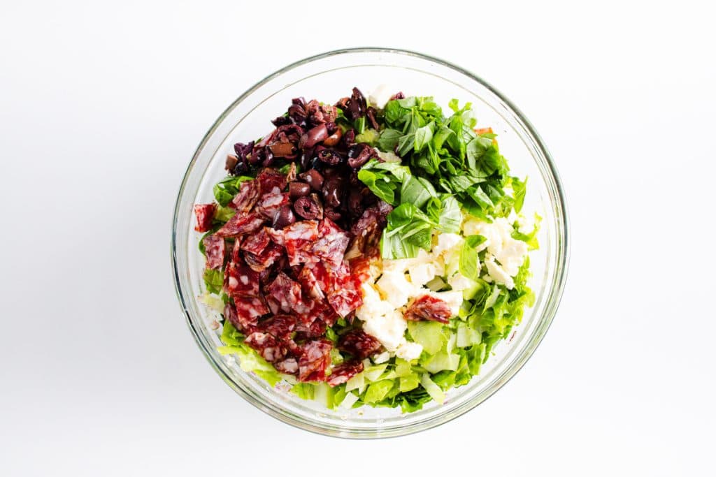 A glass bowl filled with chopped salad ingredients—lettuce, salami, olives, cheese, and fresh basil—arranged in separate sections on a white background.