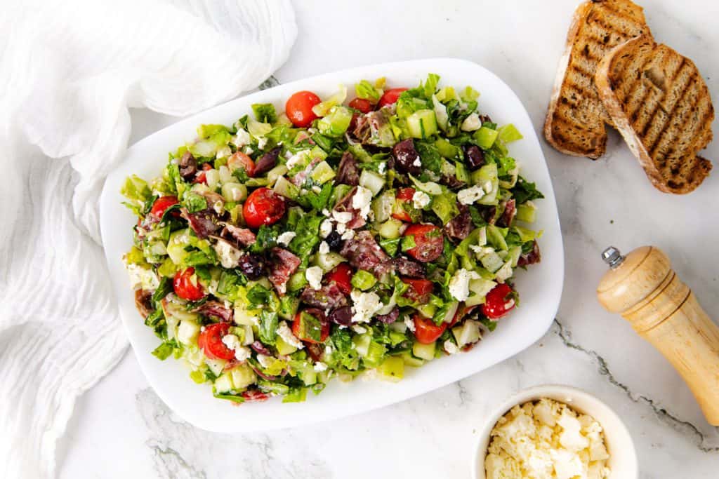 A white plate of chopped salad with crisp lettuce, juicy tomatoes, olives, and creamy feta cheese sits next to grilled bread slices, a pepper grinder, and a small bowl of crumbled feta on a marble surface.