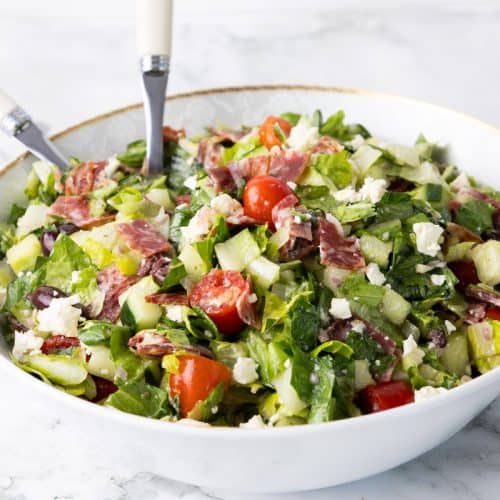 A bowl of chopped salad with lettuce, cherry tomatoes, cucumbers, olives, salami slices, and crumbled feta cheese creates a vibrant and fresh presentation, with two white-handled utensils resting inside.