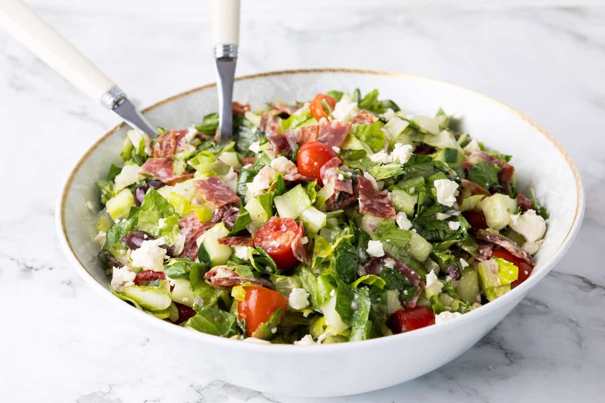 A bowl of chopped salad with lettuce, cherry tomatoes, cucumbers, olives, salami slices, and crumbled feta cheese creates a vibrant and fresh presentation, with two white-handled utensils resting inside.