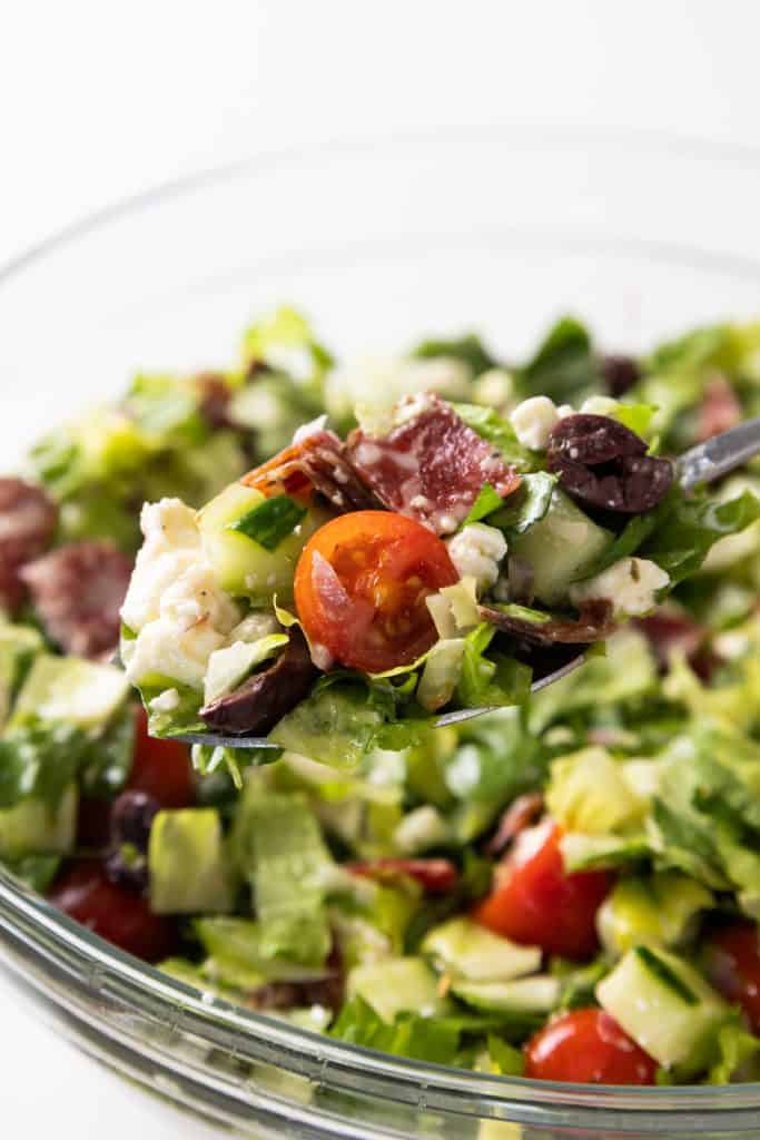 A close-up of a spoonful of chopped Greek salad with lettuce, cherry tomatoes, cucumbers, olives, feta cheese, and salami above a glass bowl filled with more vibrant chopped salad.