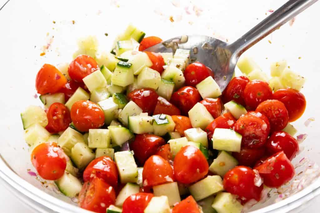 A bowl of fresh chopped salad with cucumbers, halved cherry tomatoes, diced onions, and herbs, all mixed together with a spoon.