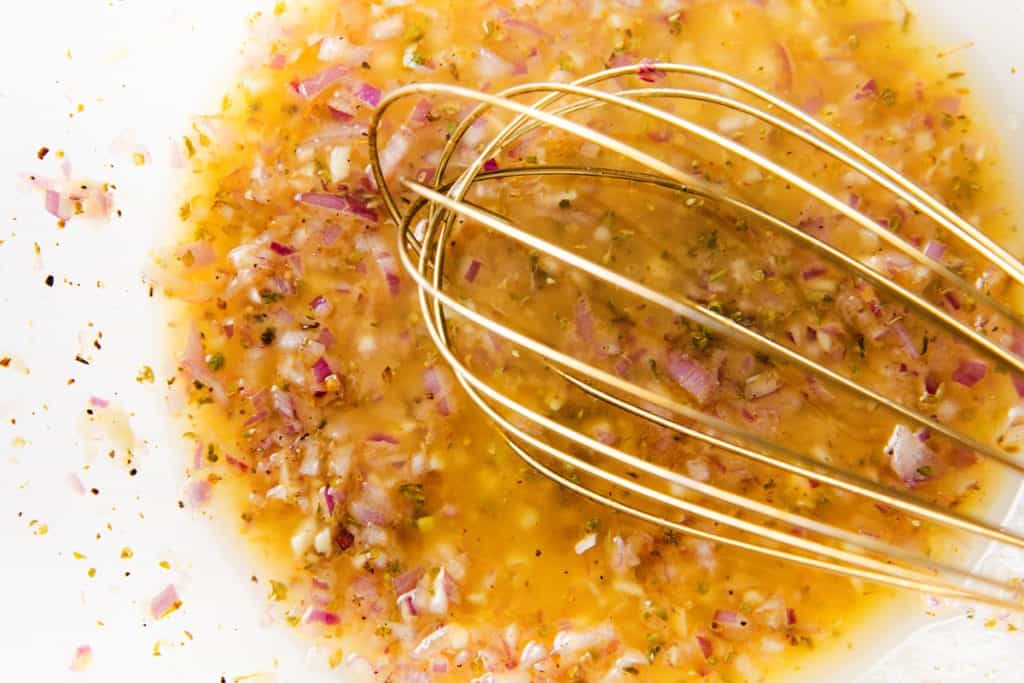 A close-up of a metal whisk mixing a marinade with chopped red onions, herbs, spices, and oil in a white bowl—perfect for drizzling over a fresh chopped salad.