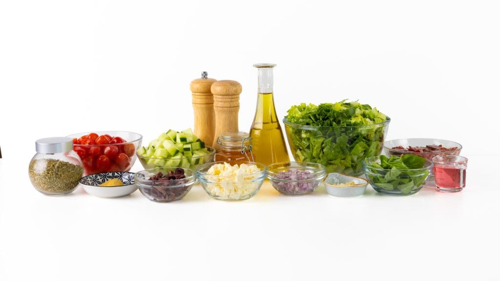 A variety of chopped salad ingredients, including lettuce, cherry tomatoes, cucumbers, cheese, olives, onions, herbs, olive oil, vinegar, and seasonings, arranged in bowls and jars on a white background.