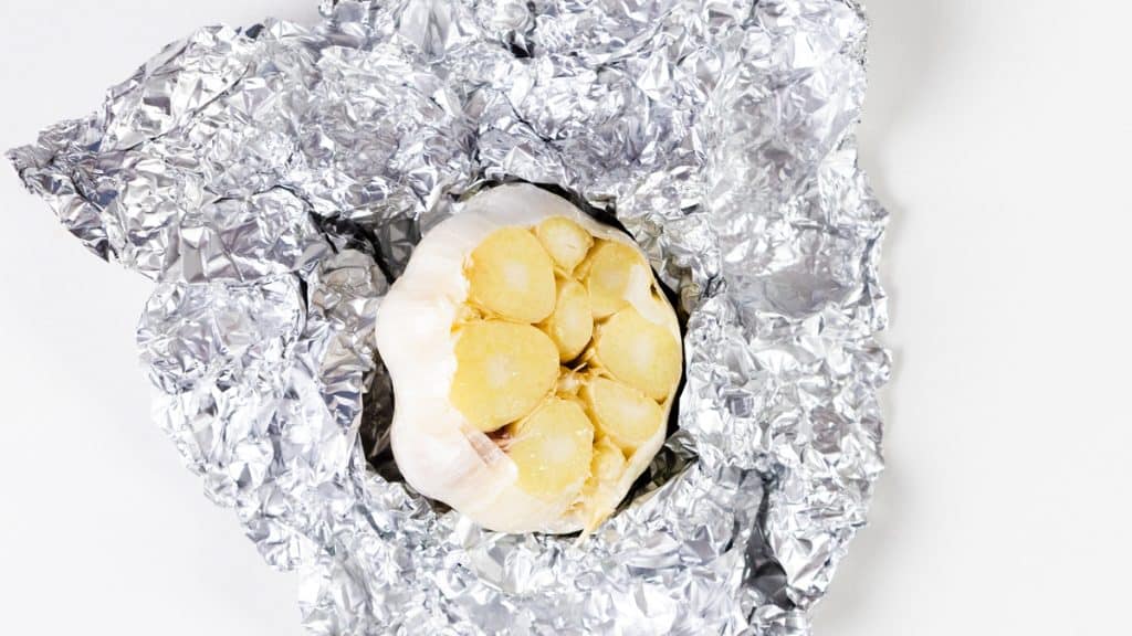 A head of garlic with the top sliced off sits on a sheet of crumpled aluminum foil, ready for roasting to infuse rich flavor into roasted summer vegetables, against a plain white background.
