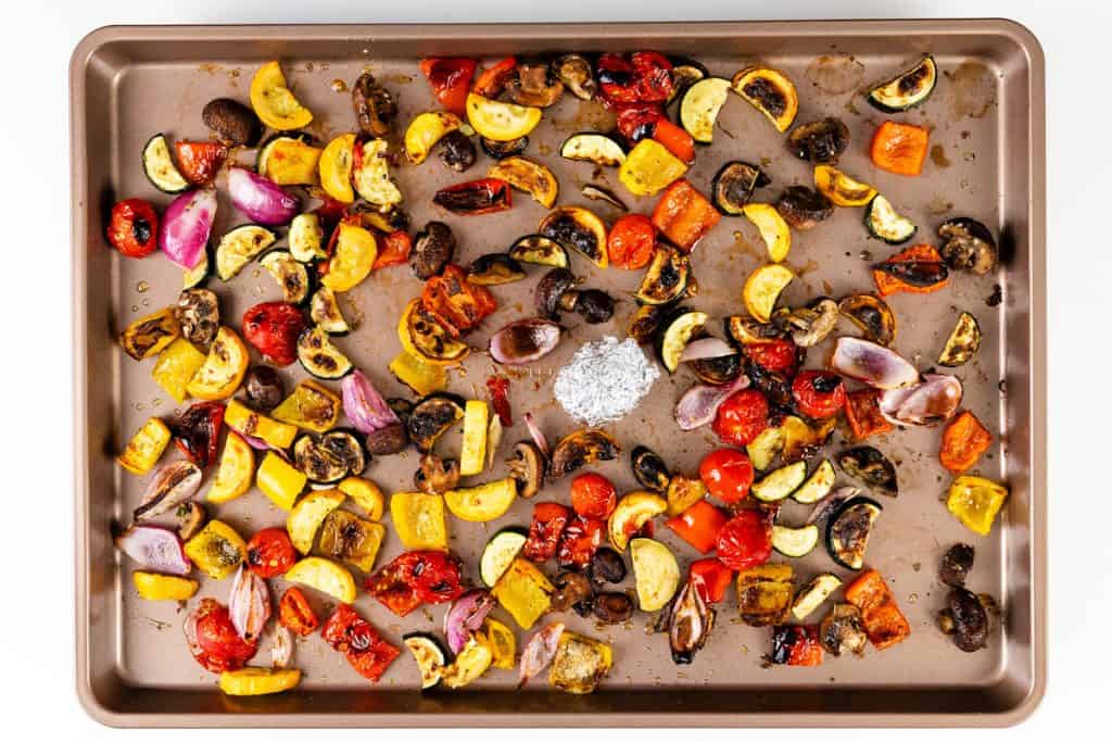 A baking sheet topped with an assortment of roasted vegetables, including summer vegetables like red onions, mushrooms, zucchini, yellow squash, and tomatoes, with a small mound of salt in the center.