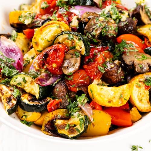 Colorful roasted vegetables, including summer vegetables like zucchini, red onion, tomatoes, bell peppers, and mushrooms, are garnished with fresh herbs and a touch of garlic-herb seasoning in a white bowl with a spoon.