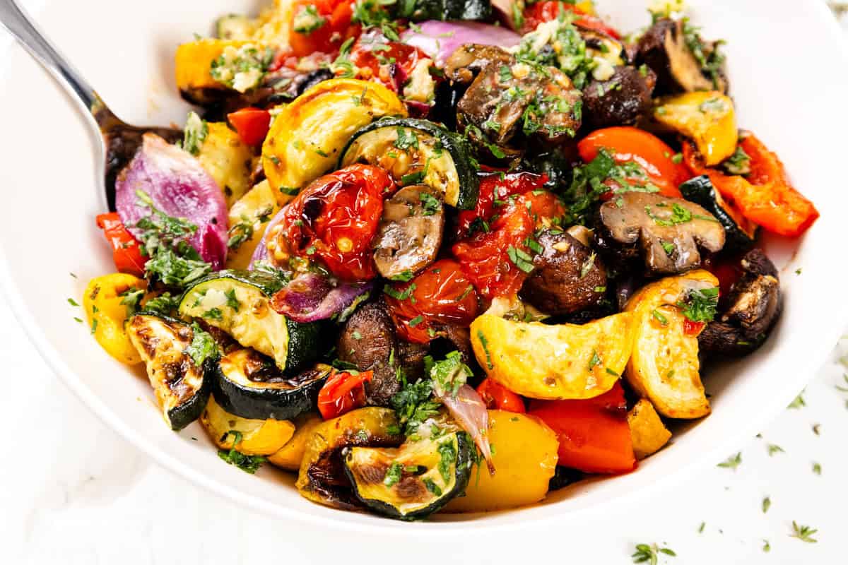 Colorful roasted vegetables, including summer vegetables like zucchini, red onion, tomatoes, bell peppers, and mushrooms, are garnished with fresh herbs and a touch of garlic-herb seasoning in a white bowl with a spoon.
