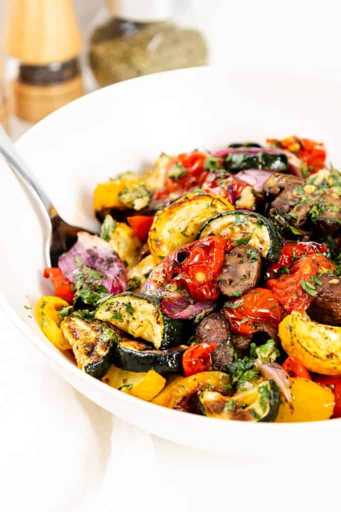 A white bowl filled with roasted summer vegetables, including zucchini, cherry tomatoes, red onions, and yellow squash, garnished with garlic-herb seasoning and fresh herbs, with a fork in the bowl.