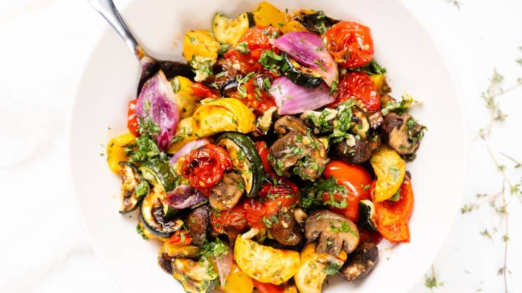 A white bowl filled with colorful roasted summer vegetables, including tomatoes, mushrooms, zucchini, red onions, and yellow squash, all garnished with chopped garlic-herb for a fresh and vibrant finish.